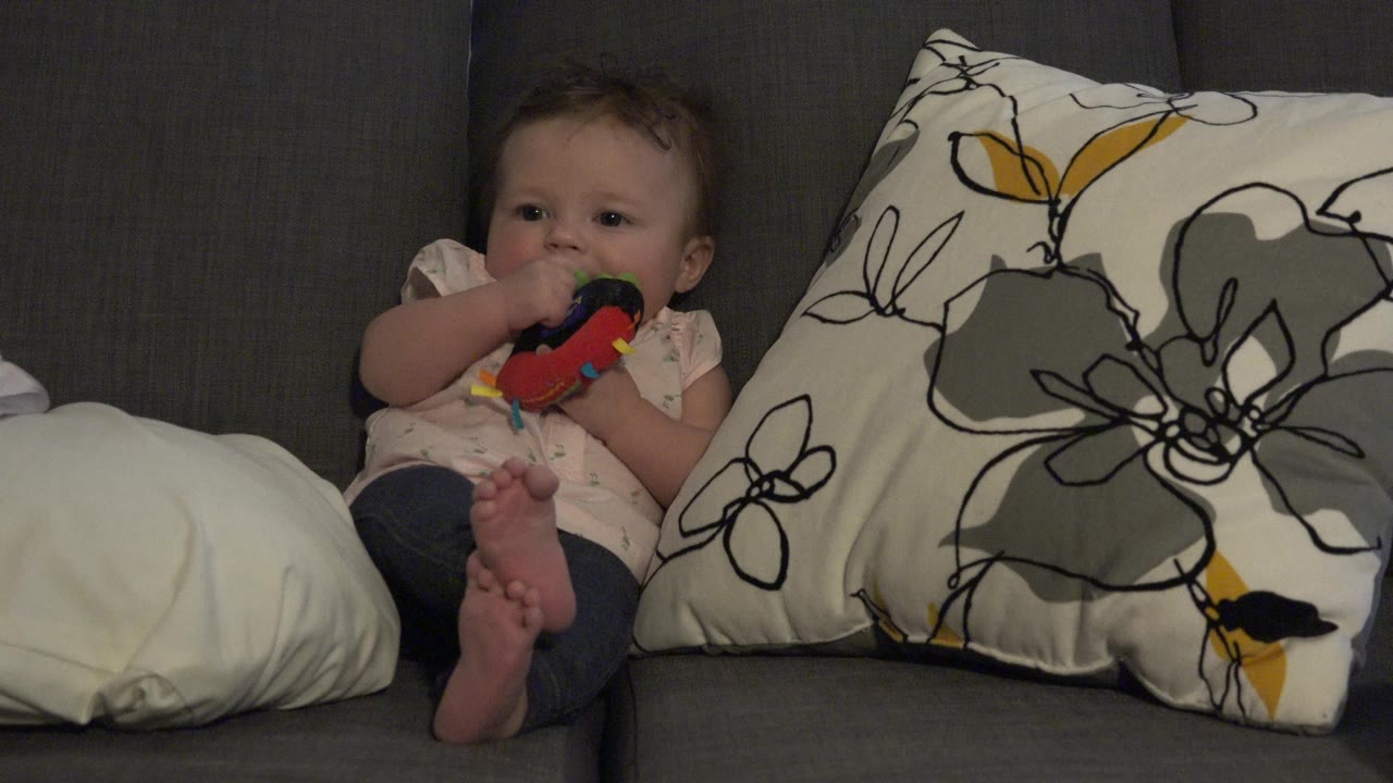 Baby seated on couch with feet forward and colorful object held at the mouth.