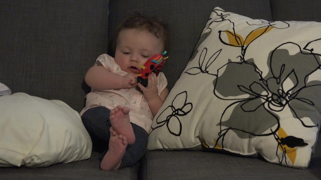 Baby girl sitting on a couch beside pillows while holding a red and black toy.