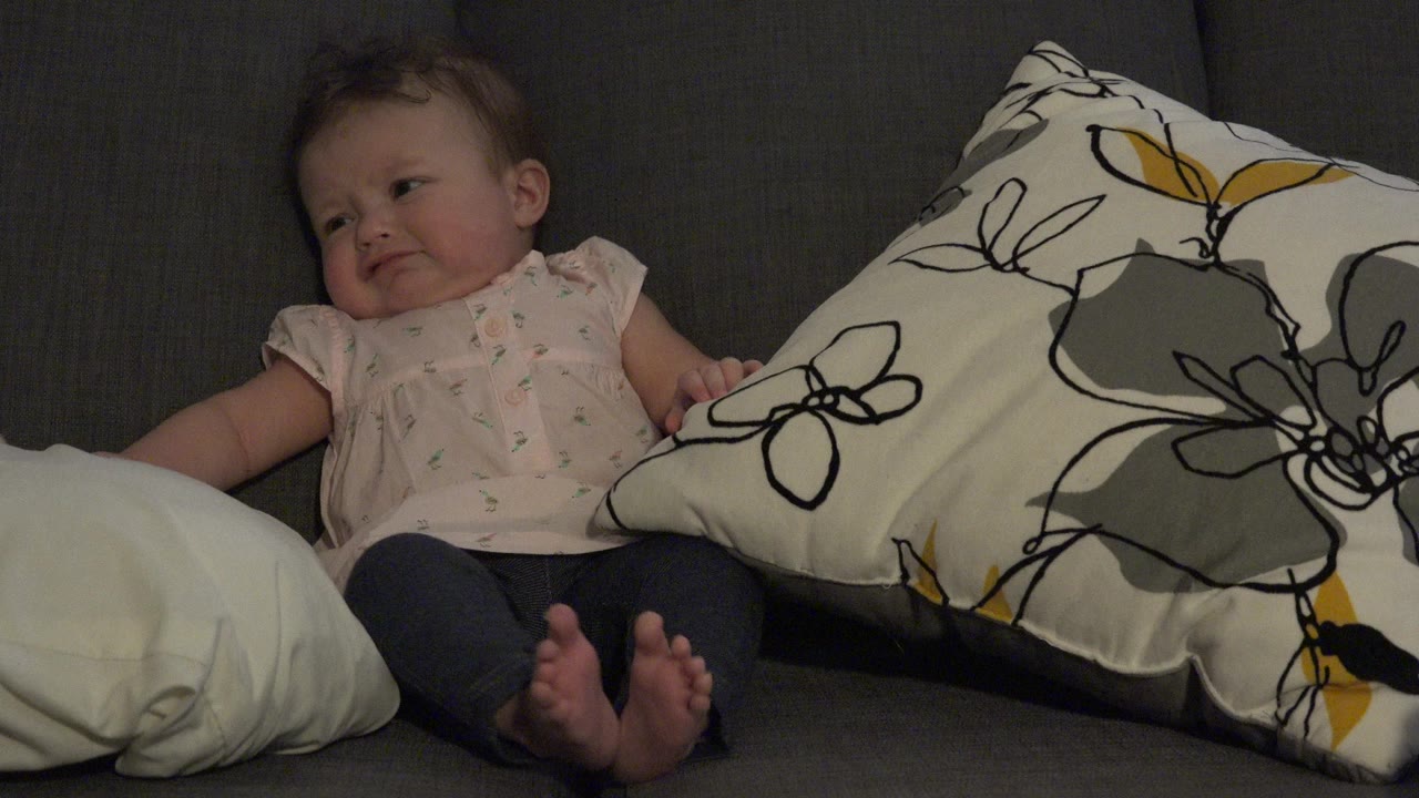 Baby seated on a gray couch beside pillows.