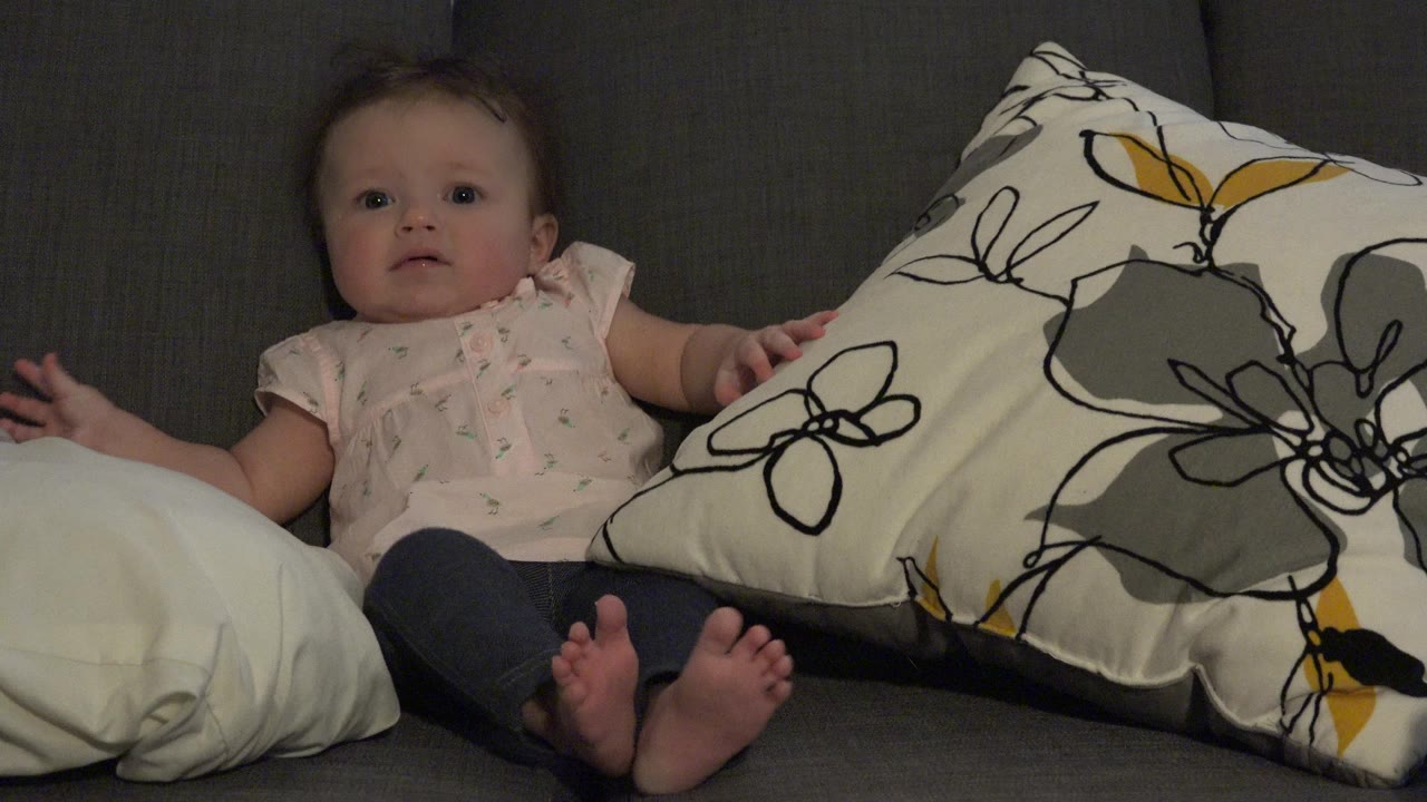 The baby remains seated on the Westminster, Colorado sofa, looking toward the camera with feet forward and the floral cushion pressed close on the right.