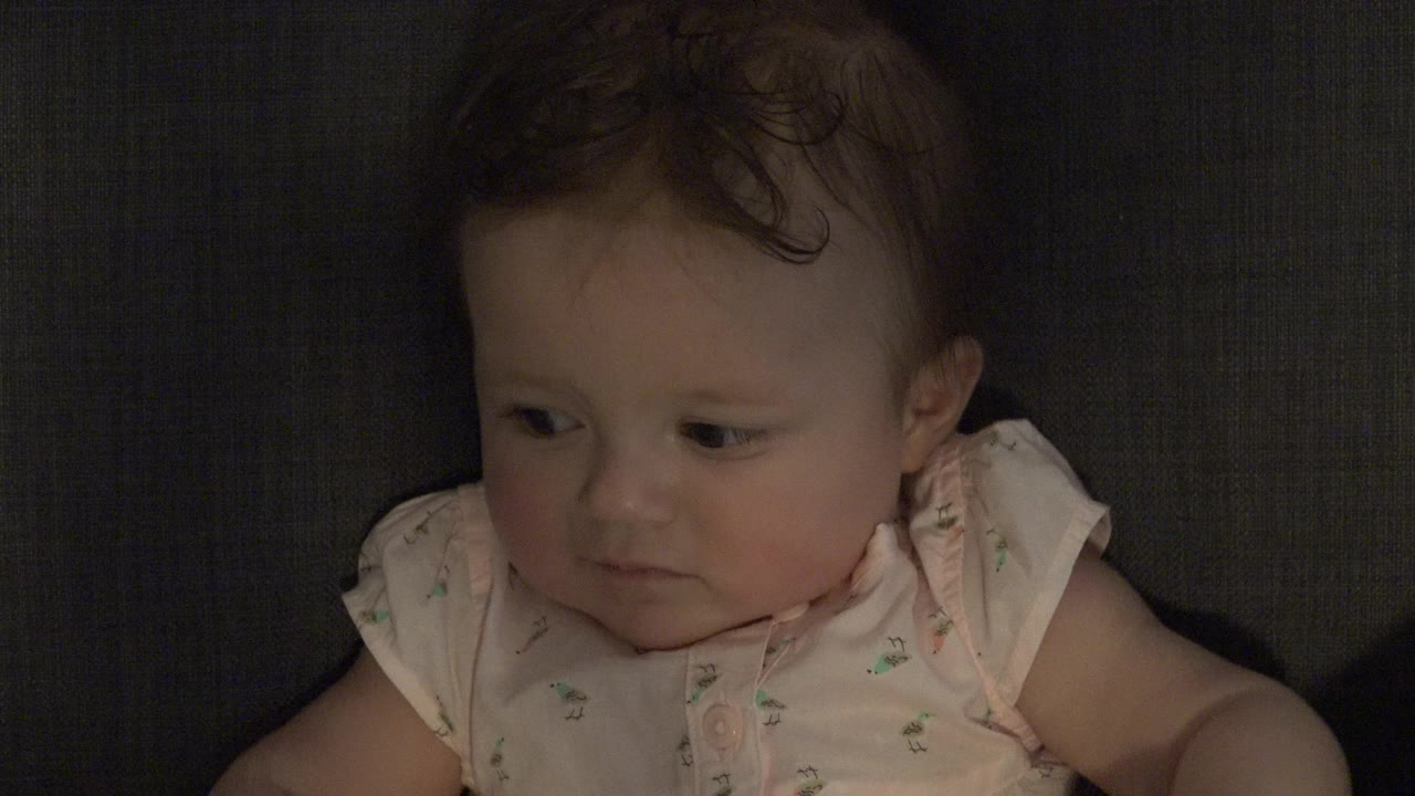 At 0:00, a baby in Westminster, Colorado sits close to the camera against a dark couch, looking down and left with a reserved expression.