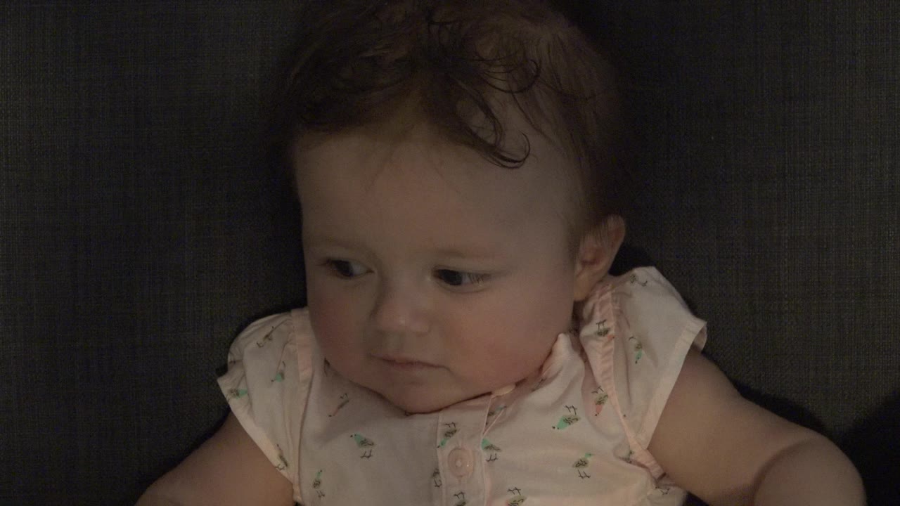 At 0:01, the baby remains seated against the dark upholstered background, eyes angled to the side and mouth relaxed.