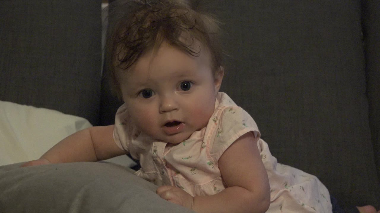 Baby girl in a pale pink outfit lies low against a gray couch cushion in Westminster, Colorado home footage, looking slightly toward the camera.