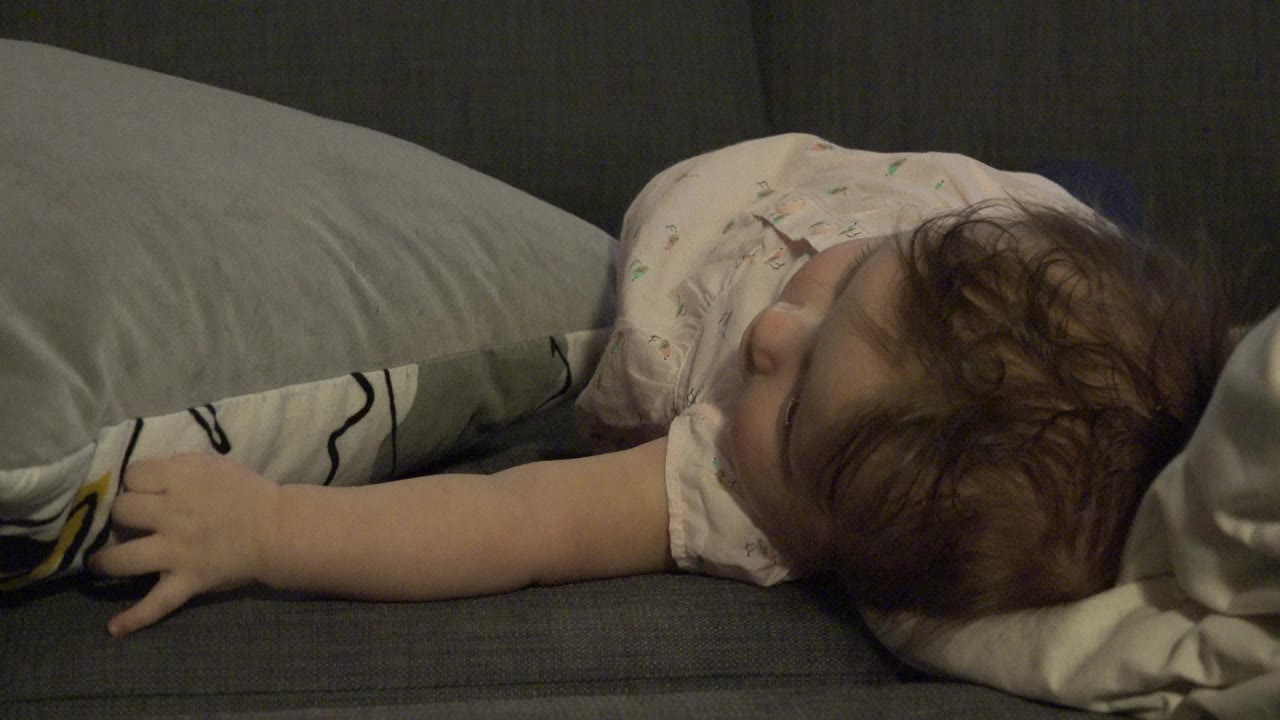 Infant lying sideways on a gray couch beside pillows.