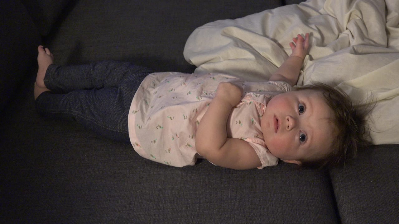 Baby lying on a gray couch beside a white blanket.
