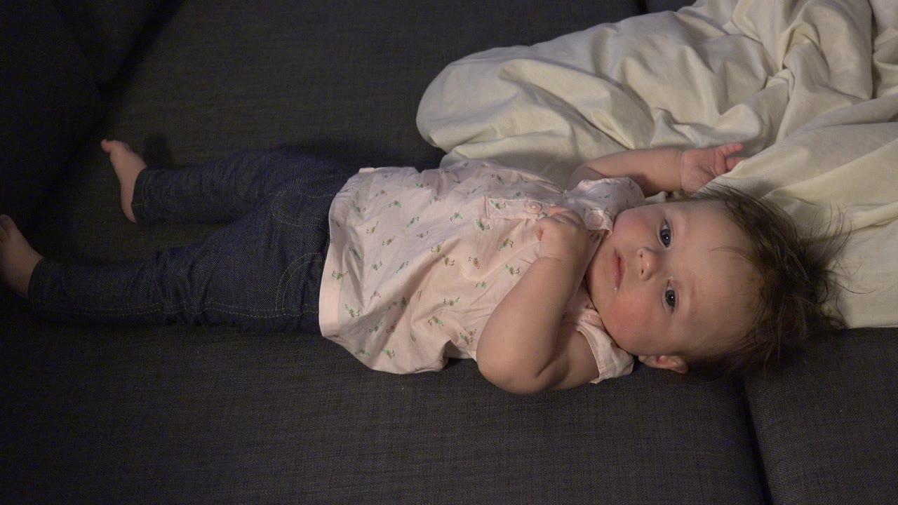 A baby lies sideways on a dark sofa in Westminster, Colorado, wearing a light patterned top and dark pants beside a white blanket.