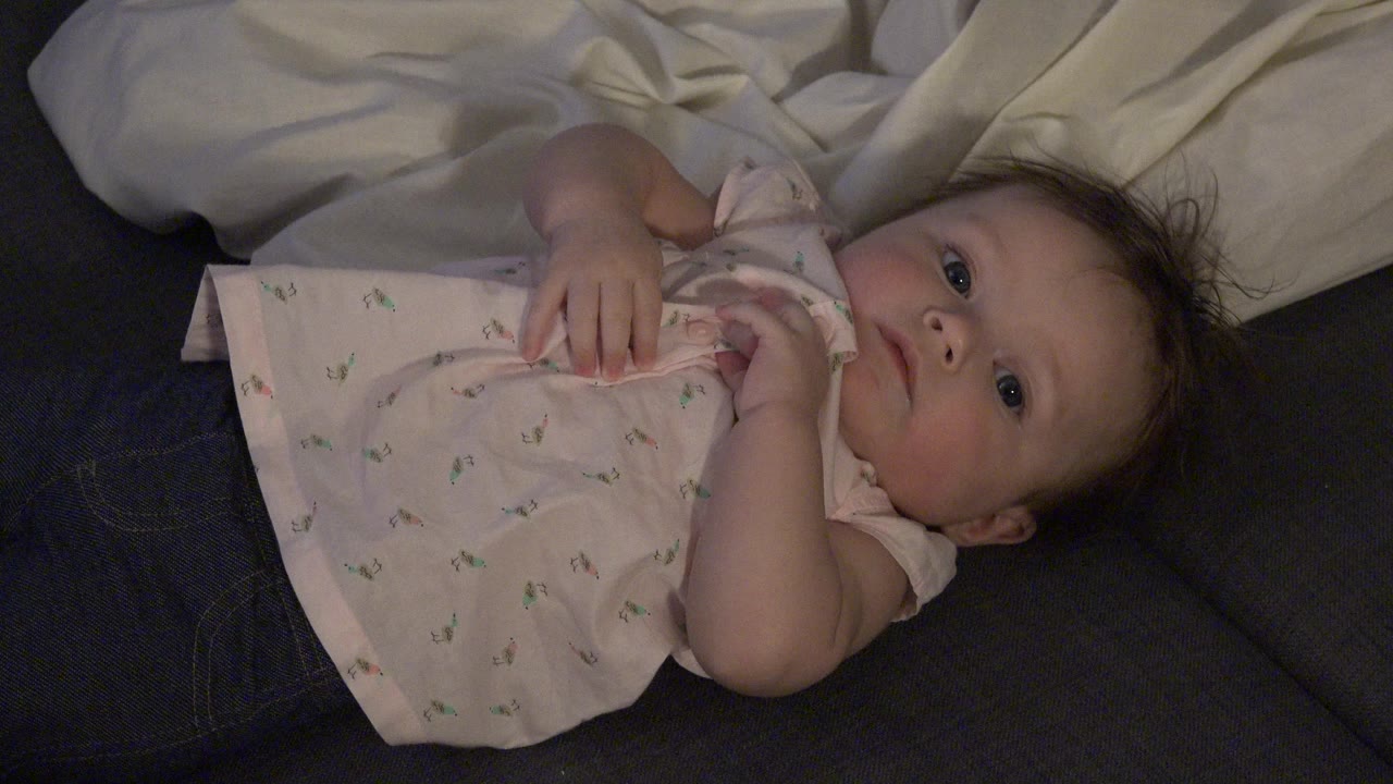 At 0:00, an infant in Westminster, Colorado lies on dark blue fabric in a pale pink patterned shirt, with white bedding behind the head and the face turned toward camera.