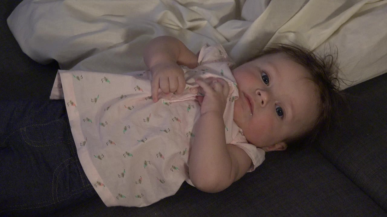 Infant with hands gathered near the shirt while lying beside white bedding.