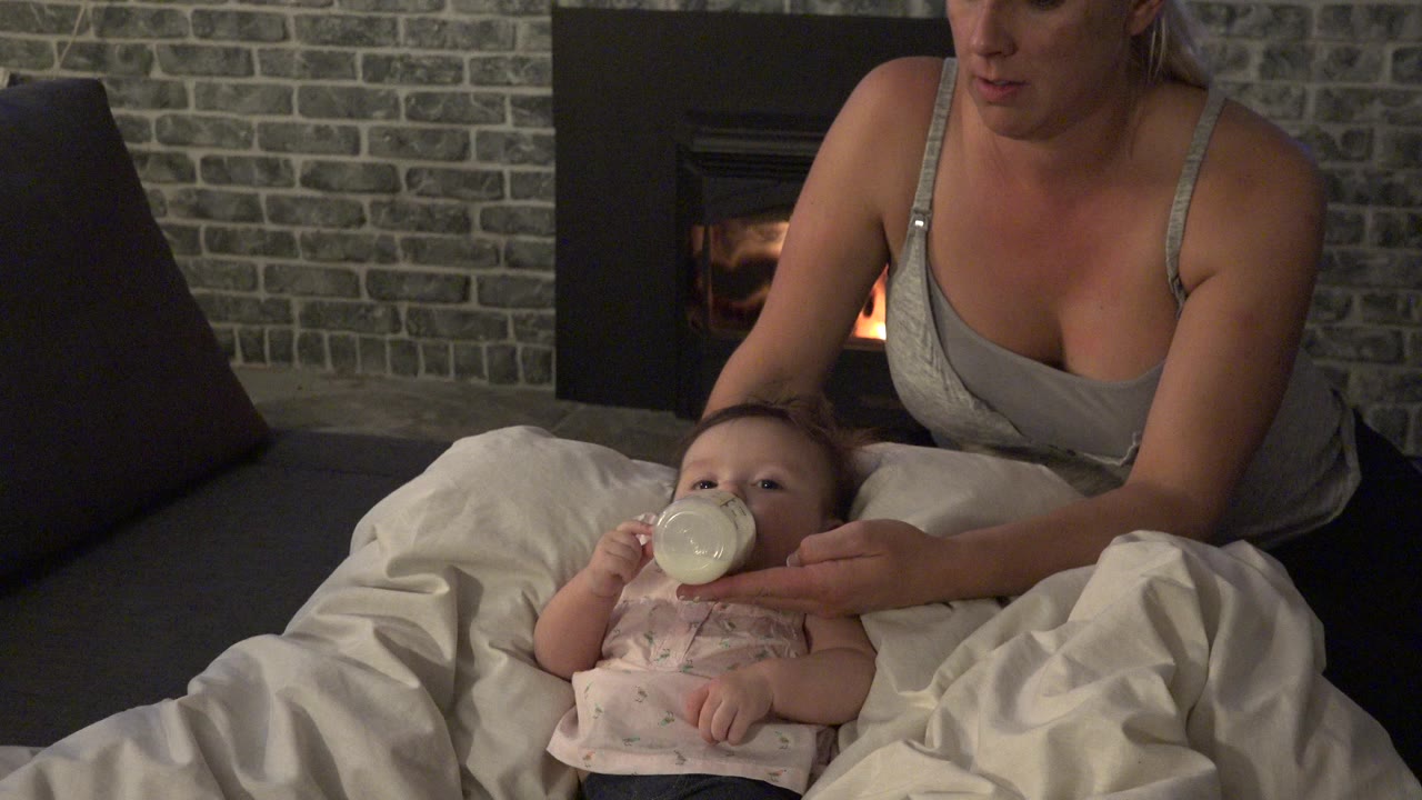 Mother holds a bottle for a baby on bedding near a fireplace.