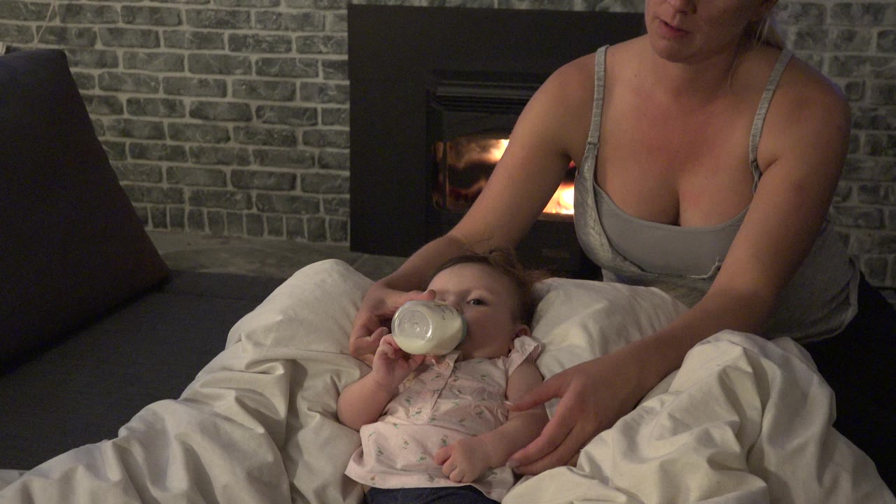 At 0:00 in Westminster, Colorado, a woman in a gray top supports a baby drinking from a bottle on white bedding with a lit fireplace and gray brick wall behind them.