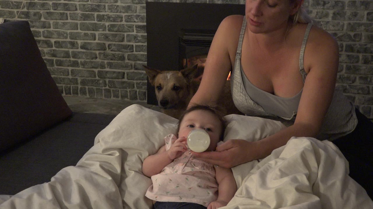 Mother supports a baby drinking from a bottle indoors while a dog looks on near a fireplace.
