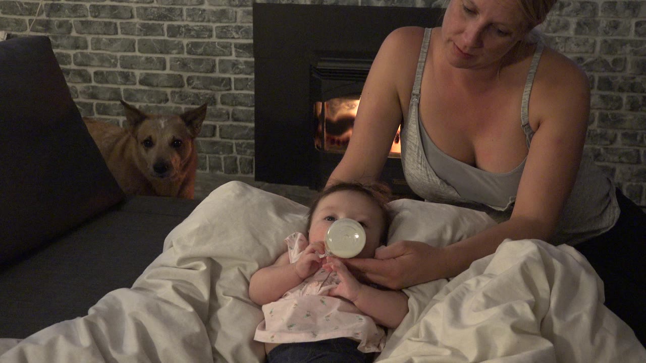 In Westminster, Colorado, the baby drinks from a bottle as the woman supports it, with the dog facing forward on the sofa and the fireplace glowing behind them.