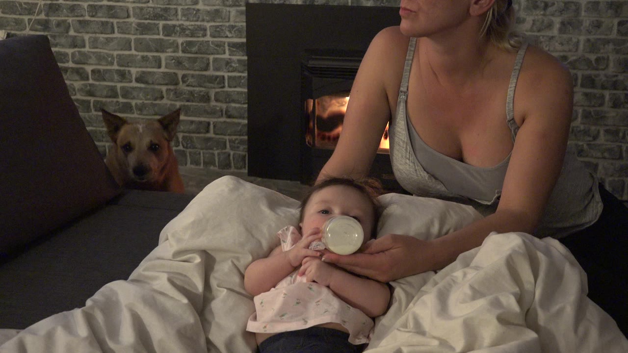 Baby drinks from a bottle beside an adult and a dog near a fireplace.
