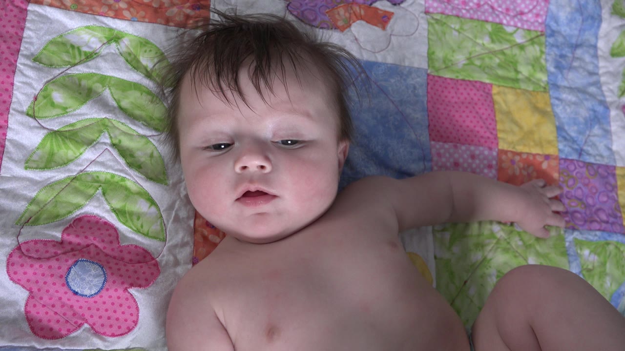 The infant remains on the pink and green quilt in Westminster, Colorado, with one arm stretched outward and eyes lowered slightly from the camera.