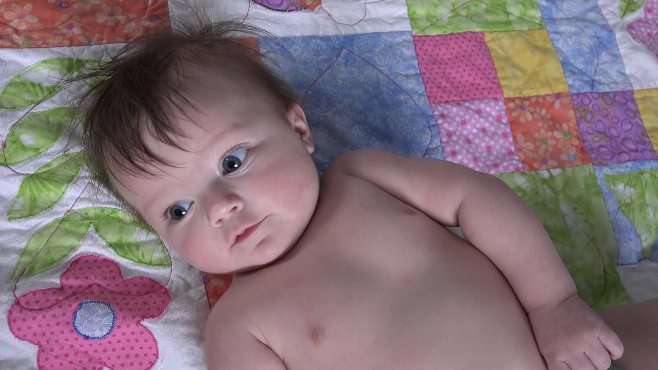 Infant girl in Westminster, Colorado remains on the colorful quilt, facing the camera with a steady neutral look and one arm bent along her side.
