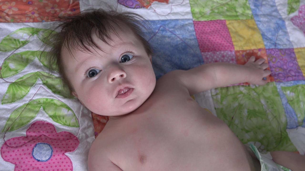 The baby remains on the patchwork blanket in Westminster, Colorado, with eyes open, lips slightly parted, and an arm reaching toward the upper right of frame.
