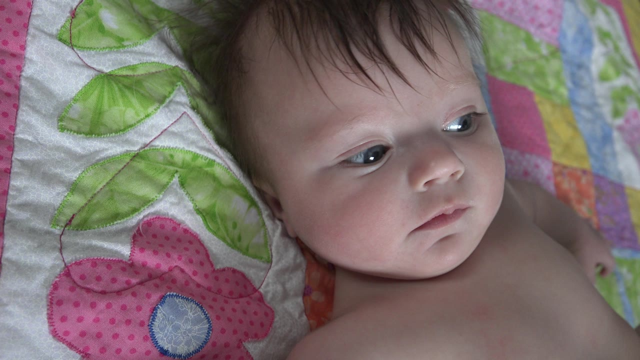 At 0:01, the baby remains on the quilt in Westminster, Colorado, with eyes turned to the side and the floral fabric filling the left side of the frame.