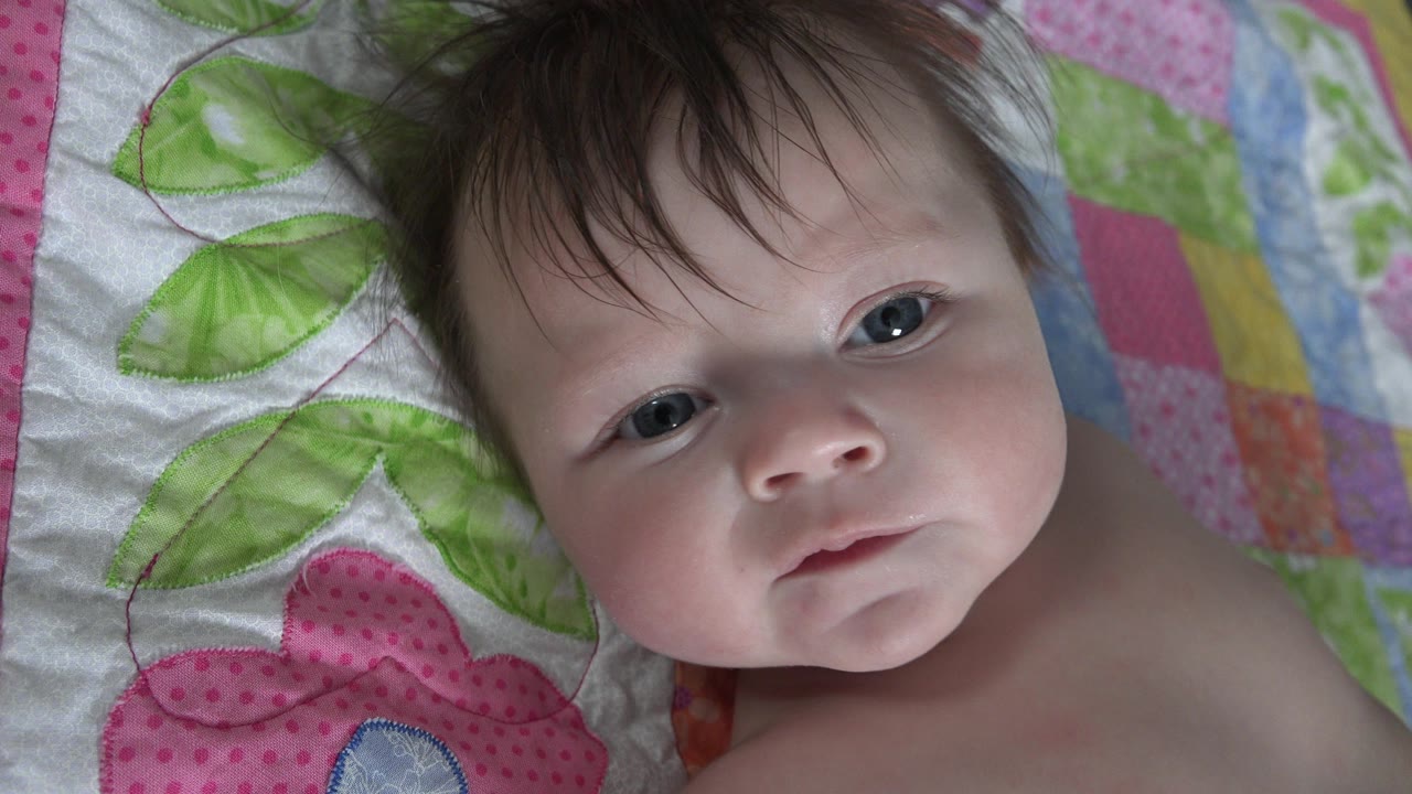 At 0:01, the infant remains centered on the colorful quilt in Westminster, Colorado, with the face turned slightly and the eyes near the lens.