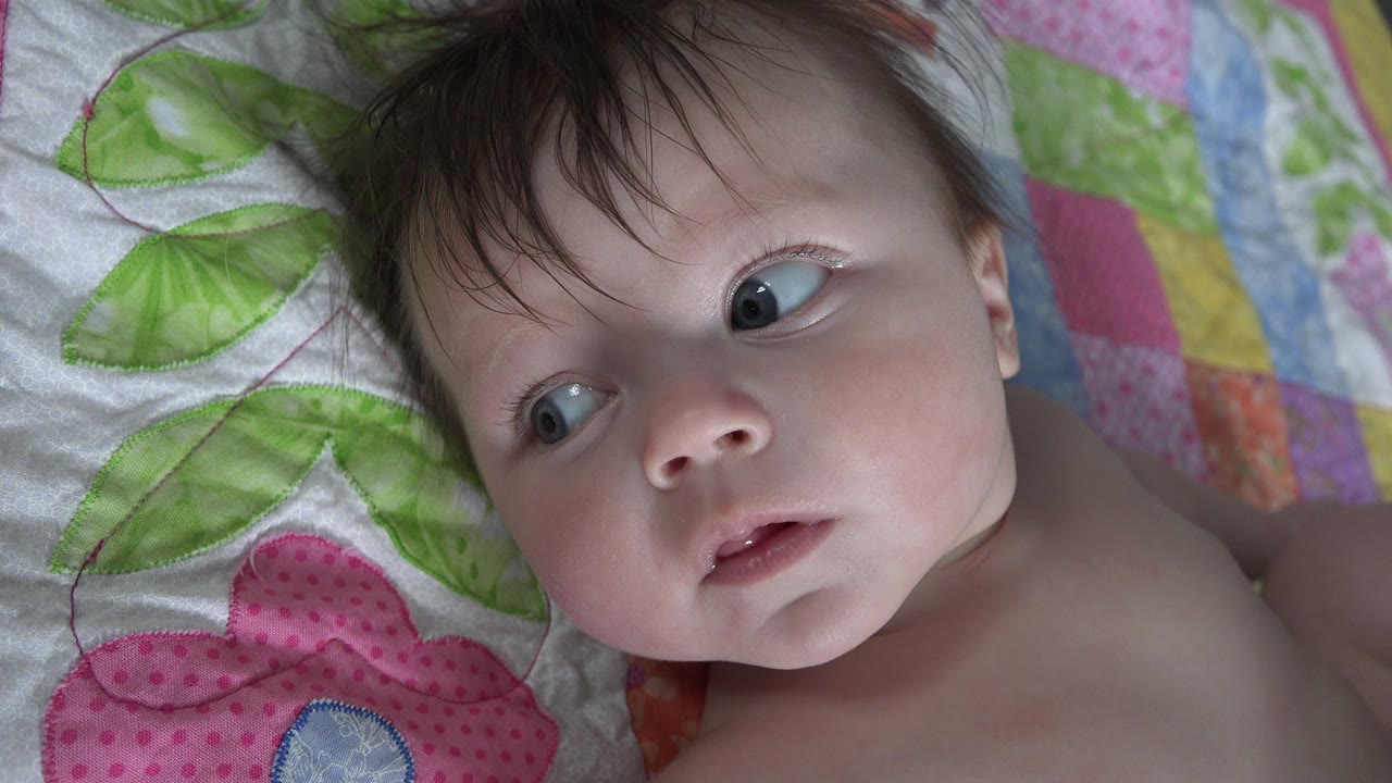At 0:00, an infant in Westminster, Colorado lies close to the camera on a patterned quilt, eyes turned toward the left side of the frame.
