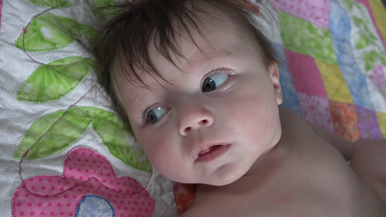 At 0:01, the infant remains on the quilt with wide eyes angled upward and left, framed tightly against green leaves and pastel fabric squares.