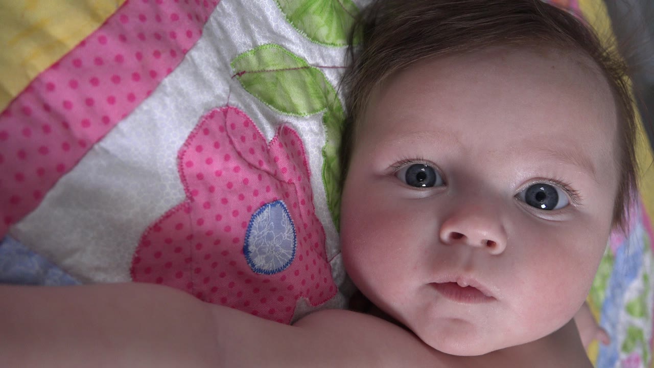 At 0:01, the infant remains nearly centered on the Westminster blanket shot, with pink polka-dot fabric and green leaf shapes visible beside her head.