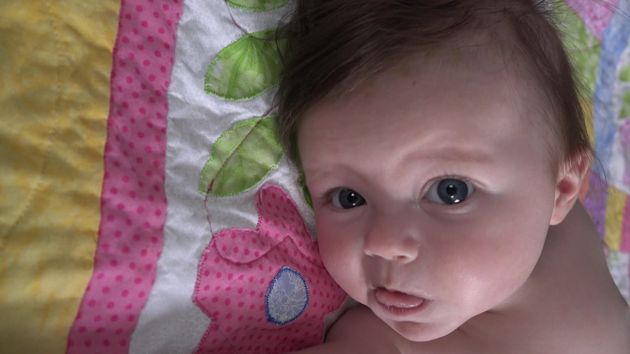 At 0:01, the baby's face remains centered against the pink and green quilt, with one eye catching more light as he looks toward the camera.