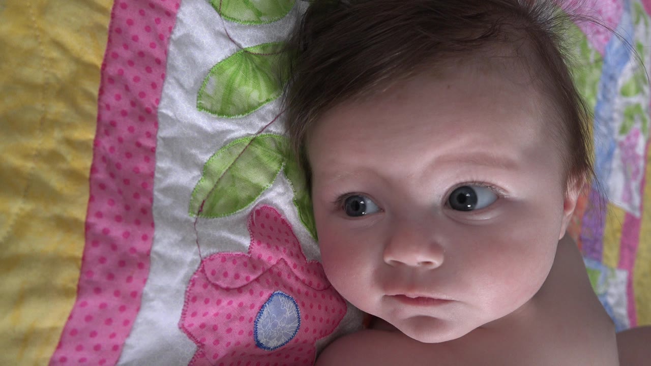 In Westminster, Colorado, a baby lies on a pink, green, yellow, and white quilt while looking left of the camera at the start of the clip.