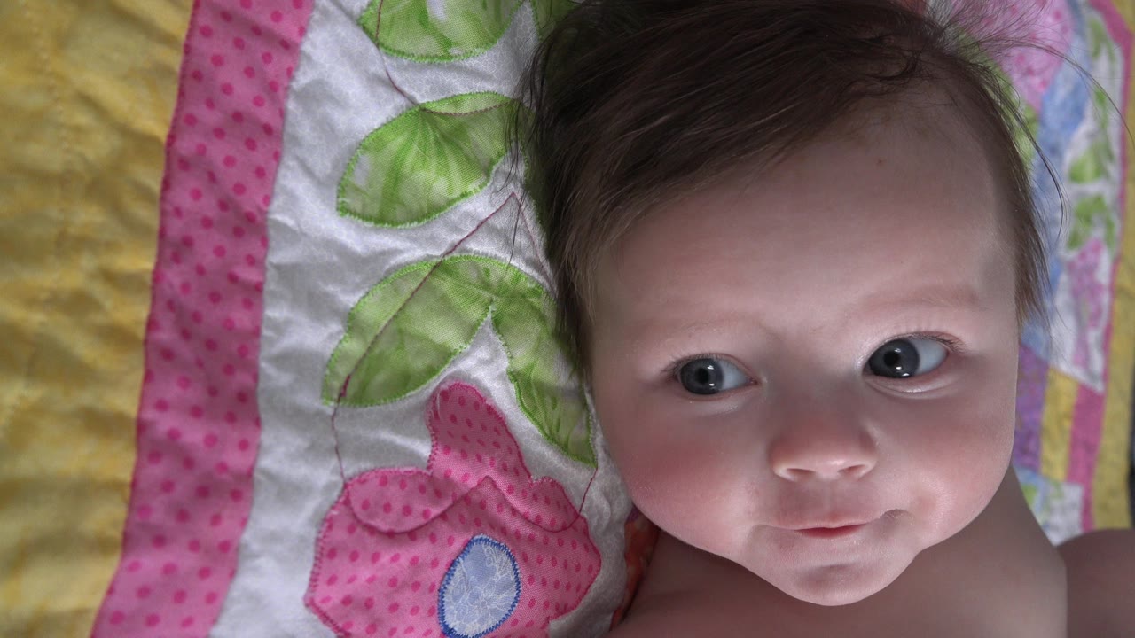 At 0:00, a baby girl in Westminster, Colorado lies close to camera on a quilt with pink trim and green leaf patterns while looking off to the side.