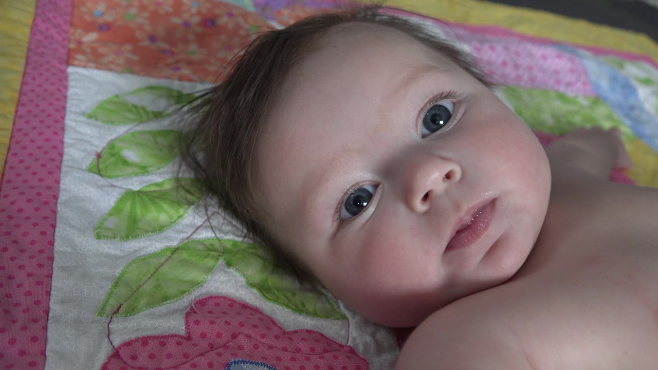 At 0:01, the baby remains framed tightly on the quilt, eyes open toward the lens with a neutral mouth and dark hair spread against the patterned fabric.