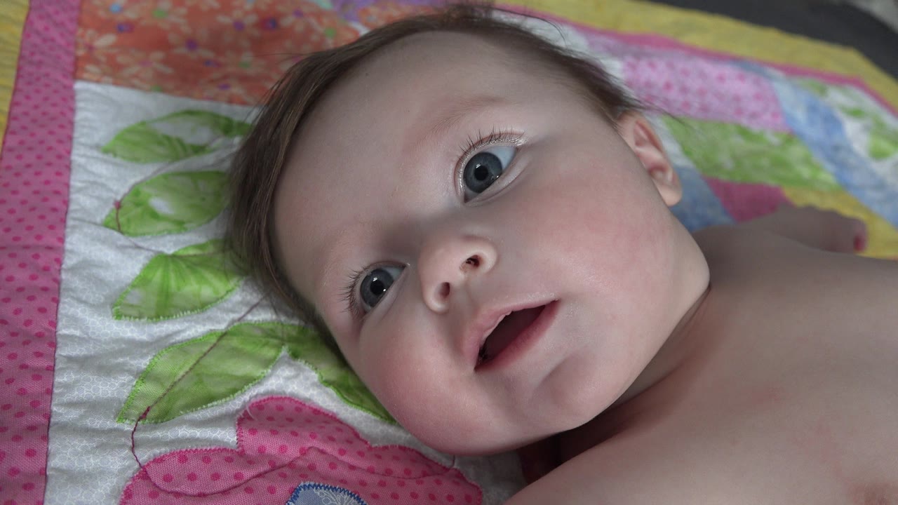 The baby remains close to camera on the colorful quilt in Westminster, Colorado, with wide blue eyes and an open mouth during the babbling moment.