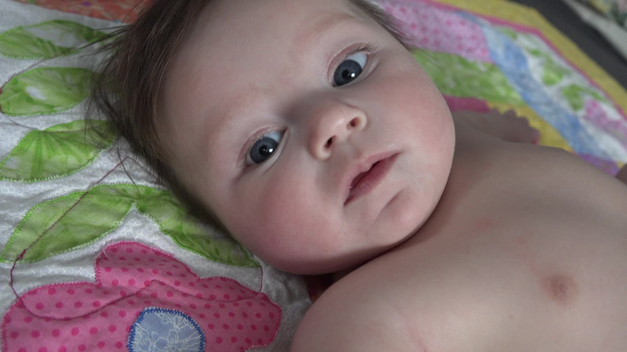 At 0:01, the baby remains on the colorful bedding with wide eyes, parted lips, and bare shoulder visible in the close frame.