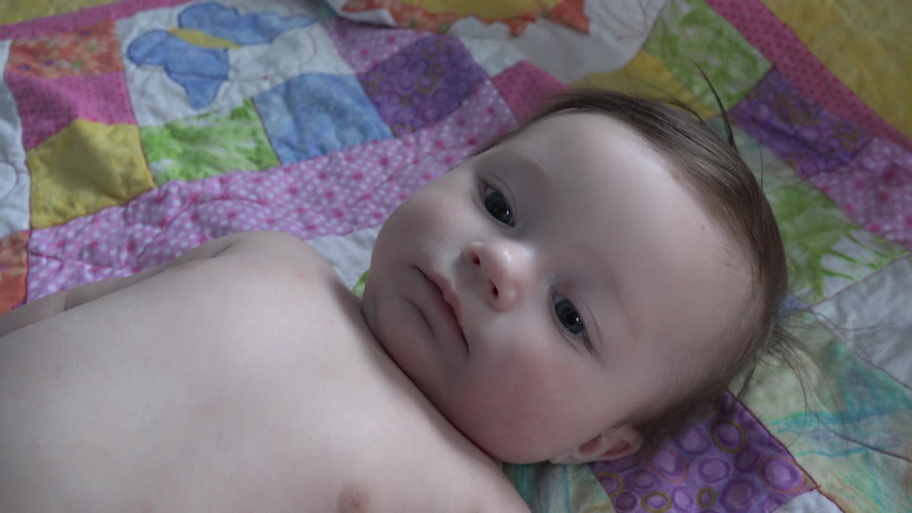 At 0:01, the baby remains on the Westminster bed quilt with eyes open and head resting against purple and pink fabric patches.