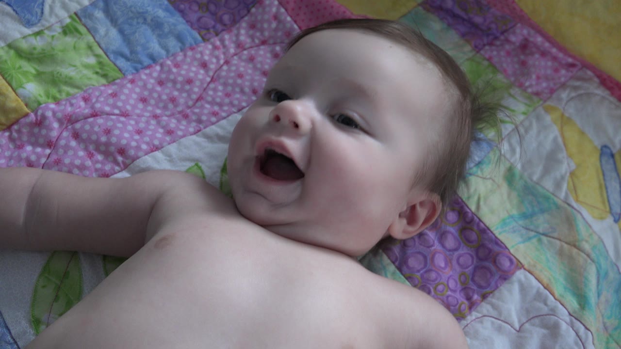 At 0:00, a baby in Westminster, Colorado lies close to camera on a pink and purple patchwork quilt with an open-mouthed smile.