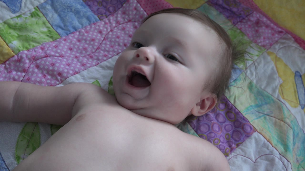At 0:01, the baby remains on the colorful quilt with the mouth open wider and the head turned slightly toward the side.
