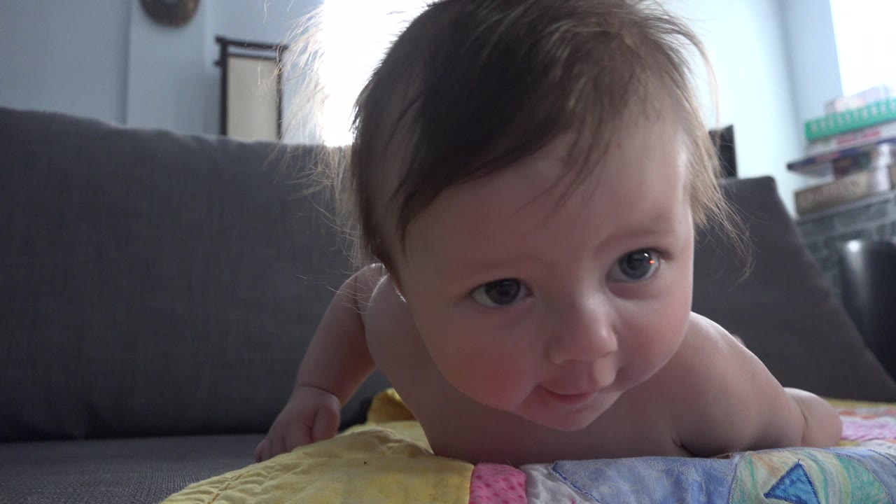 At 0:01, the baby remains propped on the colorful mat, with her face turned forward and the gray couch filling most of the background in the Westminster home interior.