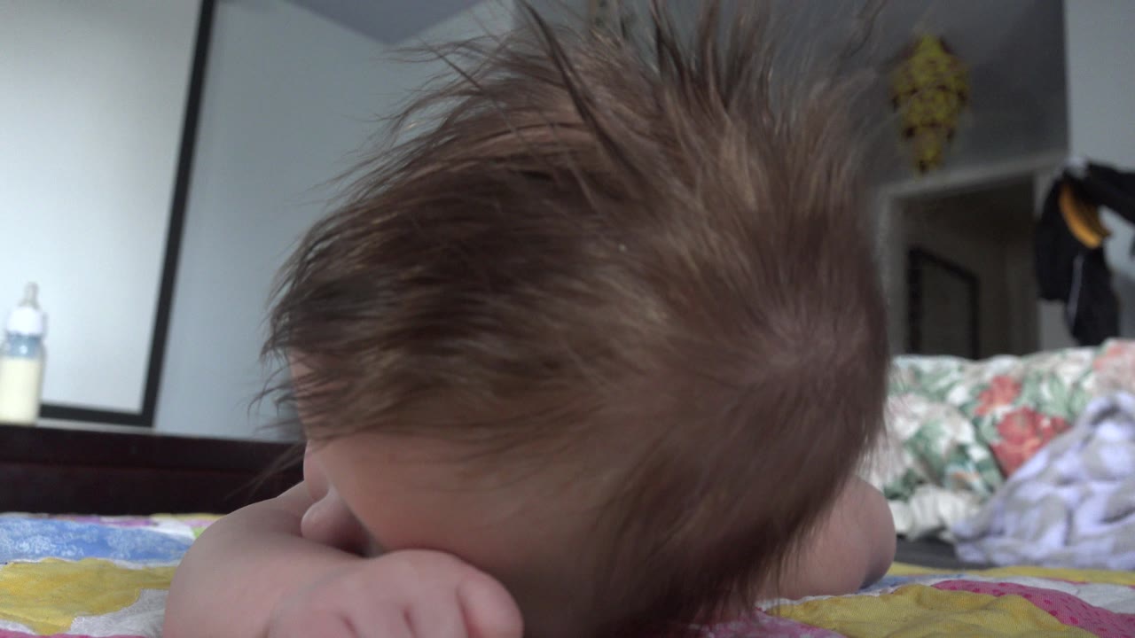 At 0:00 in Westminster, Colorado, a baby rests face-down on a patterned bedspread with tousled hair prominent and bedroom furniture blurred behind.