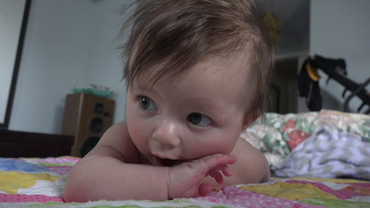 At 0:01, the baby remains close to the quilt with one hand in the mouth, eyes turned sideways, and the bedroom background softly out of focus.