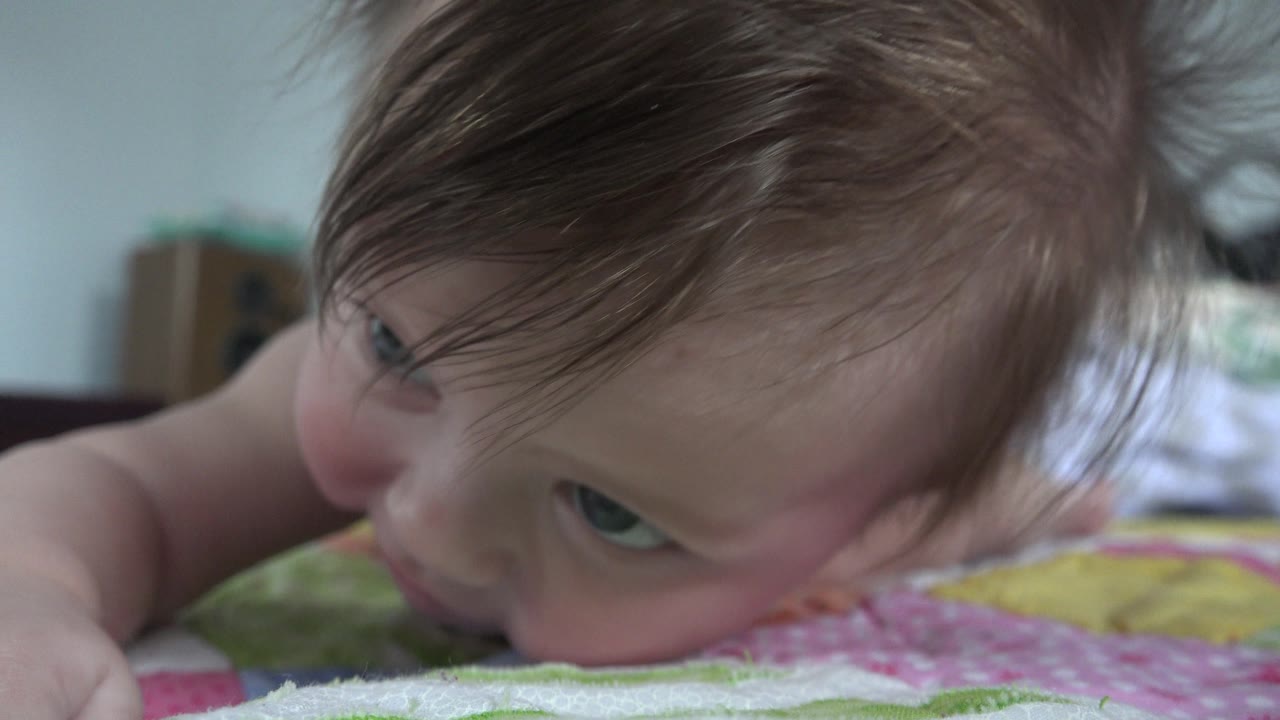 Baby lifts face from the bed and looks sideways near the camera.