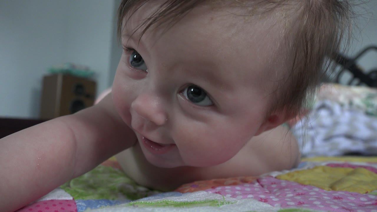A baby lies close to the camera on a colorful bedspread in a Westminster, Colorado home interior, looking upward with one arm extended in the foreground.