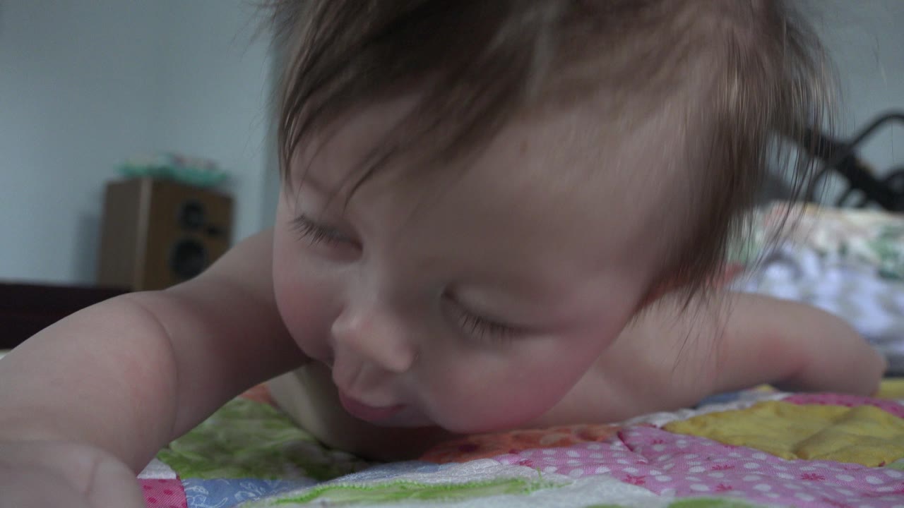 At 0:01 in Westminster, Colorado, the baby remains face down on the quilt, head turned slightly down, with soft bedding and a pale wall behind.