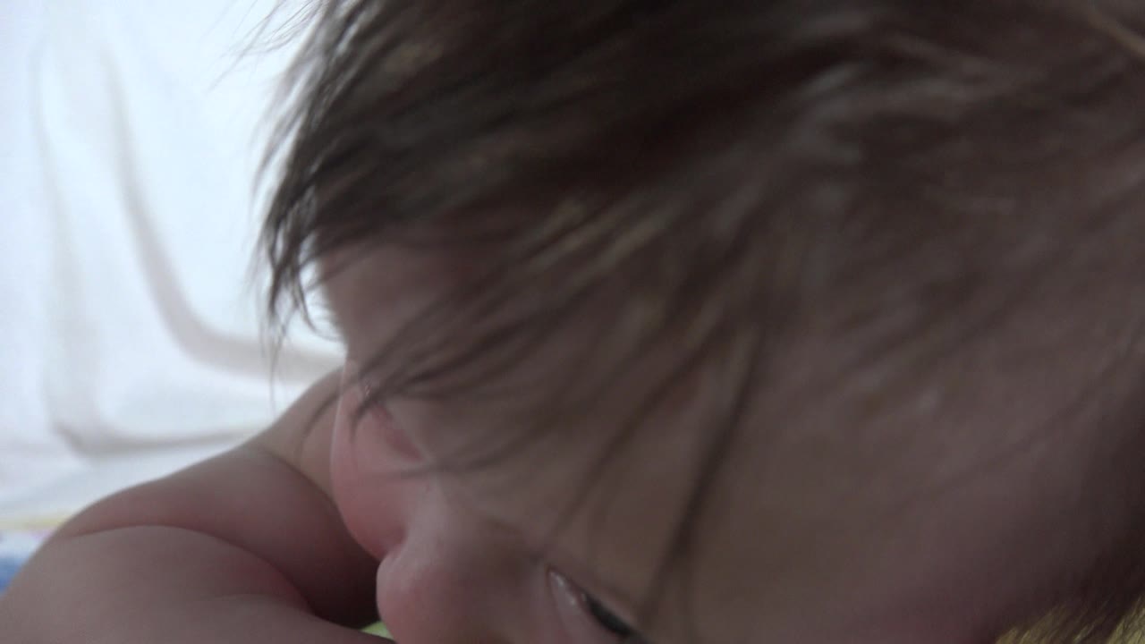 The infant remains in extreme close-up in Westminster, Colorado, with the head angled downward, hair strands across the face, and pale bedding softly blurred behind.