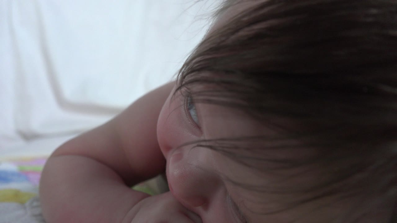 At 0:00, a baby lies on a bed in Westminster, Colorado, with dark hair across the forehead and cheek, white bedding behind the head, and a patterned blanket at the lower edge.