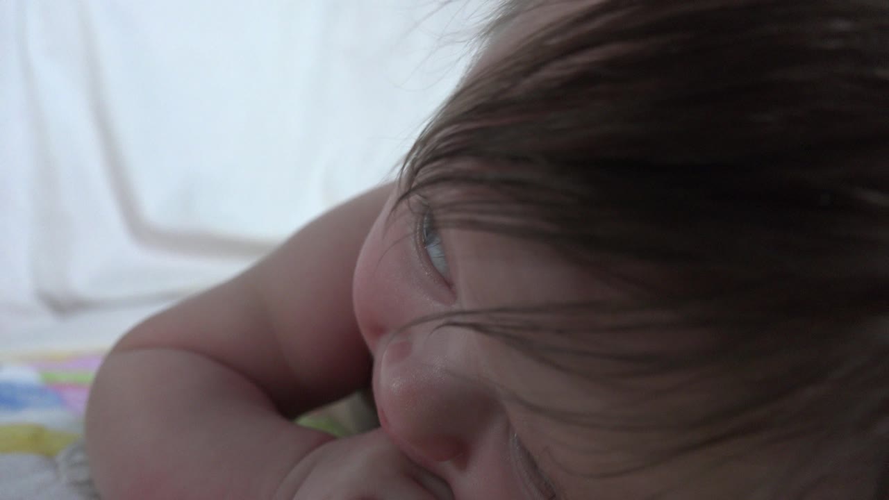 At 0:01, the infant remains in a tight bed-level close-up in Westminster, Colorado, with the face angled downward, hair covering one eye area, and pale bedding filling the background.