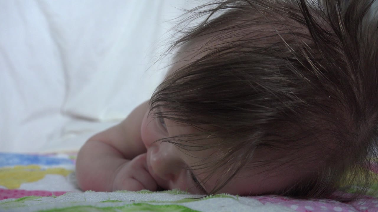 Very young infant lying face-down on a patterned blanket with long brown hair.