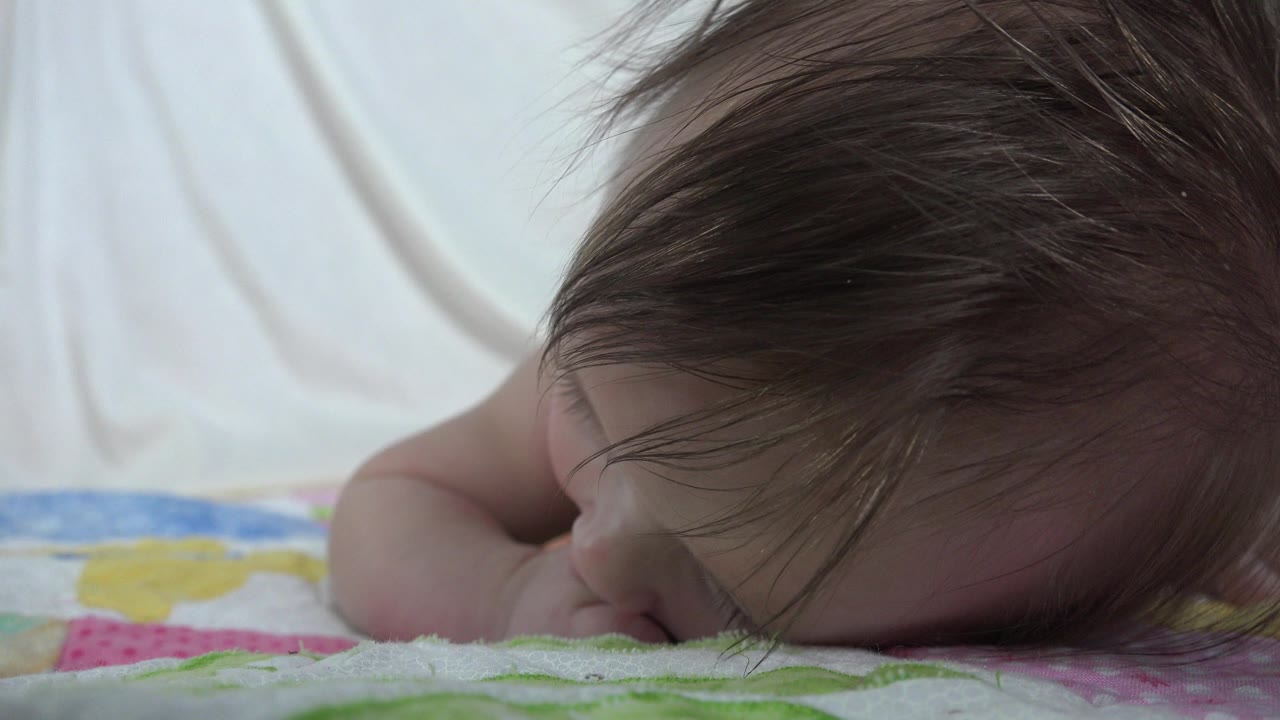 At 0:01, the infant remains low against the quilt in Westminster, Colorado, with one arm tucked forward and the child's face mostly turned down.