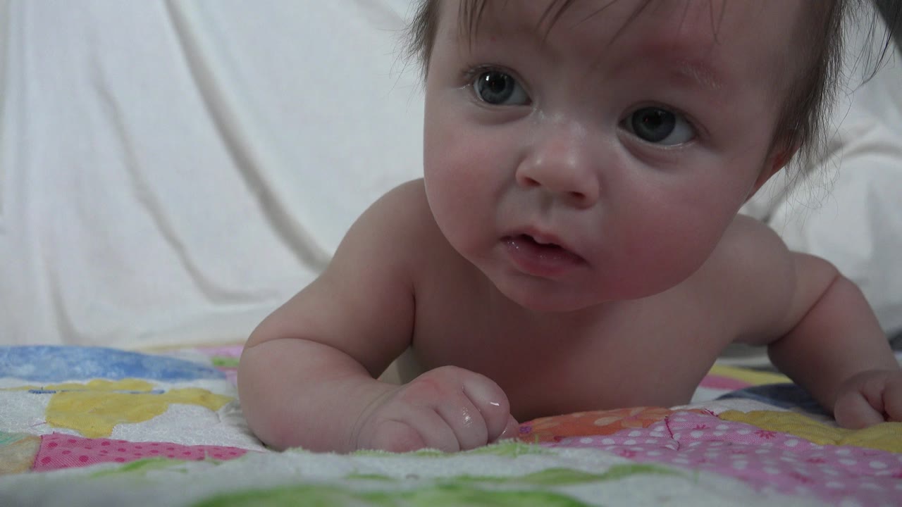 A close view shows the baby propped on his arms on the patterned blanket, facing near the camera with damp hair and soft indoor light.