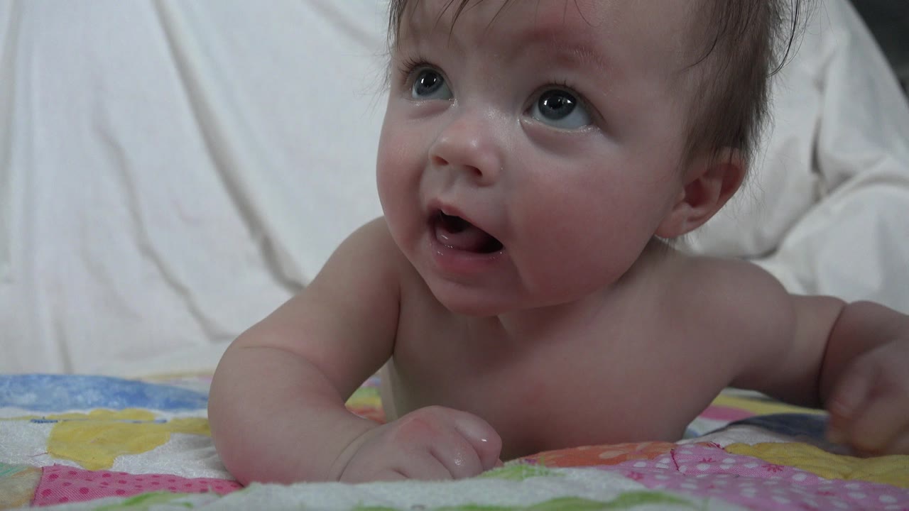 At 0:01, the baby remains propped on forearms on the bed, mouth open and eyes raised toward something above the camera.