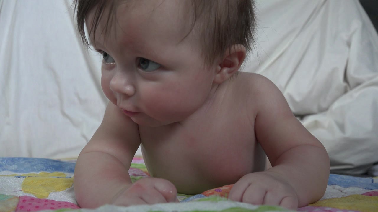 A white baby in Westminster, Colorado lies on her stomach on a colorful blanket with white bedding behind her, looking off to the left at the start of the clip.