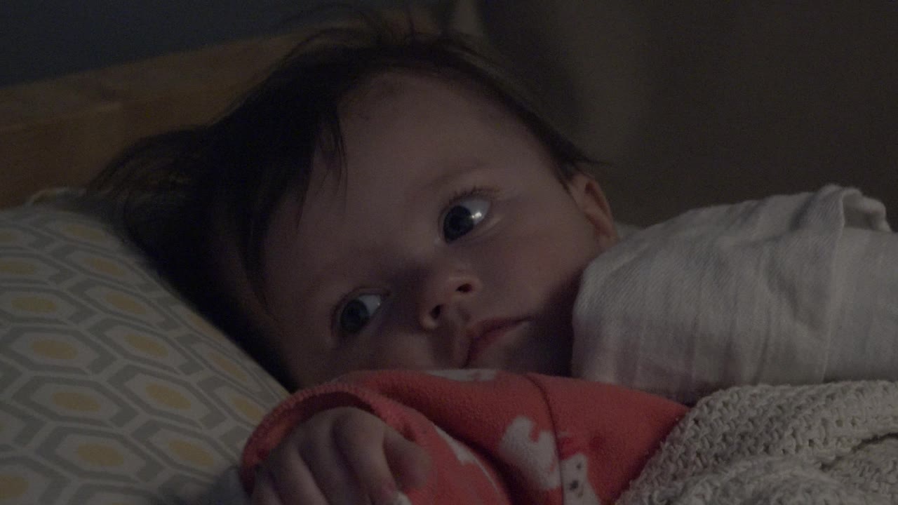 A close view shows the baby looking off to the side from the pillow, with dark hair spread against the bedding and a white blanket at right.