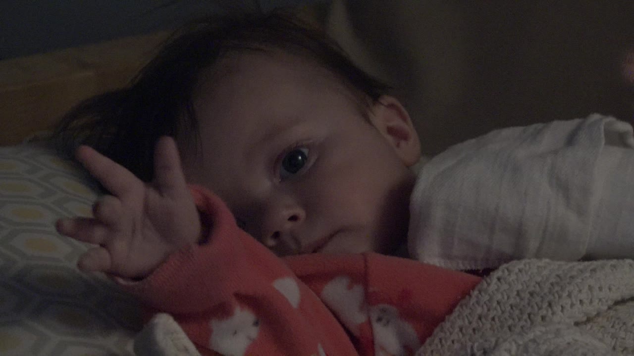 In Westminster, Colorado, a baby in a red sleeper lies on patterned pillows in dim indoor light with one hand lifted beside the face.